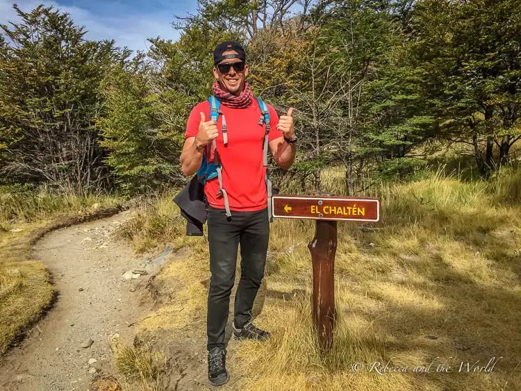 A smiling hiker - the author's husband - wearing a red shirt, sunglasses, and a backpack gives a thumbs-up beside a wooden signpost indicating the direction to El Chaltén. The trail behind him leads through a forested area.