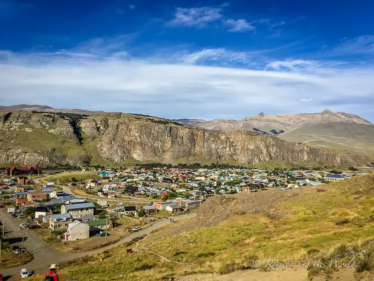 A panoramic view of El Chaltén village, nestled at the foot of a large hill with colorful houses scattered across the landscape, framed by distant mountains under a clear blue sky. The lure of El Chalten's hiking trails draw trekkers from all around the world. Located in Argentine Patagonia, this small town is perfect for whiling away a few days and getting adventurous. Just don't get lost on one of the El Chalten hiking trails! Read on for what to expect, how to get to El Chalten and where to stay. | #argentina #patagonia #elchalten #elchaltenthingstodo #elchaltenhiking #elchaltenpatagonia #travelguide #hiking