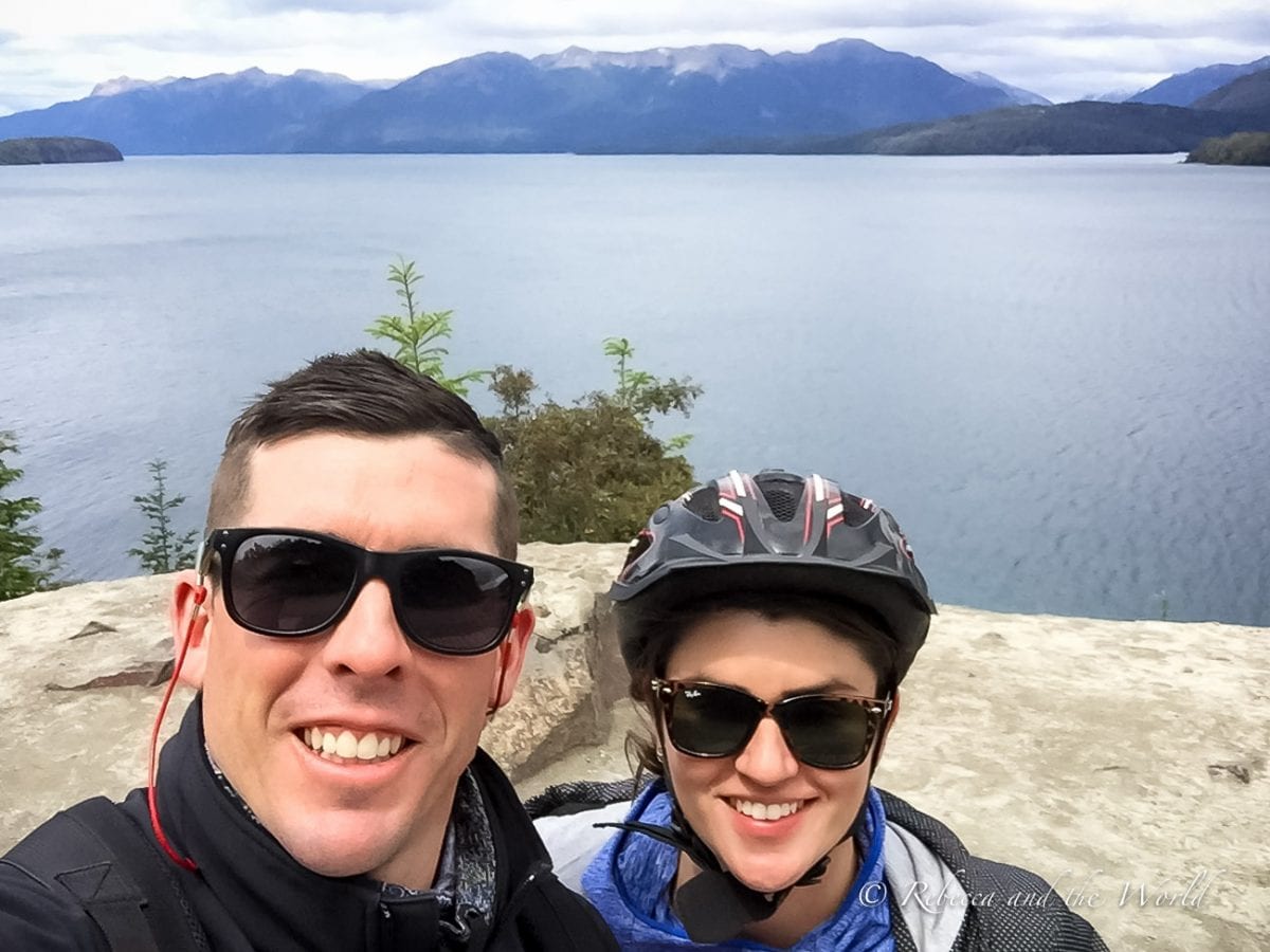 A selfie of a smiling man and woman (the author and her husband) wearing sunglasses and helmets, with a lake and mountains in the background. Try biking the Ruta de los Siete Lagos route over 3 days - it's a great adventure to have in Argentina!