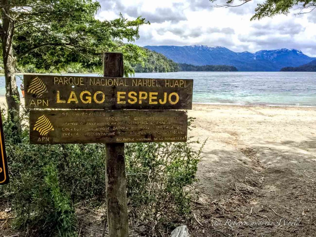 A wooden sign for "PARQUE NACIONAL NAHUEL HUAPI LAGO ESPEJO" beside a clear lake with a sandy shore, backed by forested mountains under a partly cloudy sky. Lago Espejo is one of the lakes you can visit while traversing the Ruta de los 7 Lagos in Argentina.