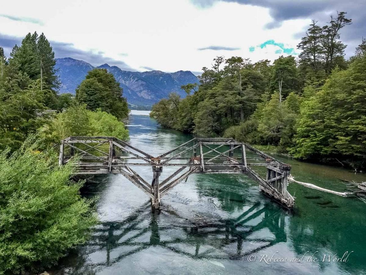 An aerial view of a derelict bridge over a clear, calm river surrounded by dense green forest. Mountains loom in the distance under a cloudy sky. There are more than a dozen lakes along the Ruta de los Siete Lagos in Argentina, along with picturesque rivers and streams.