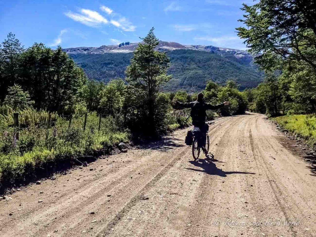 A cyclist (the author's husband) on a dirt road with arms raised in joy, facing away from the camera. Lush greenery surrounds the road, with a mountain in the distance under a clear blue sky. Biking is a great way to visit the 7 Lakes Route in Argentina.