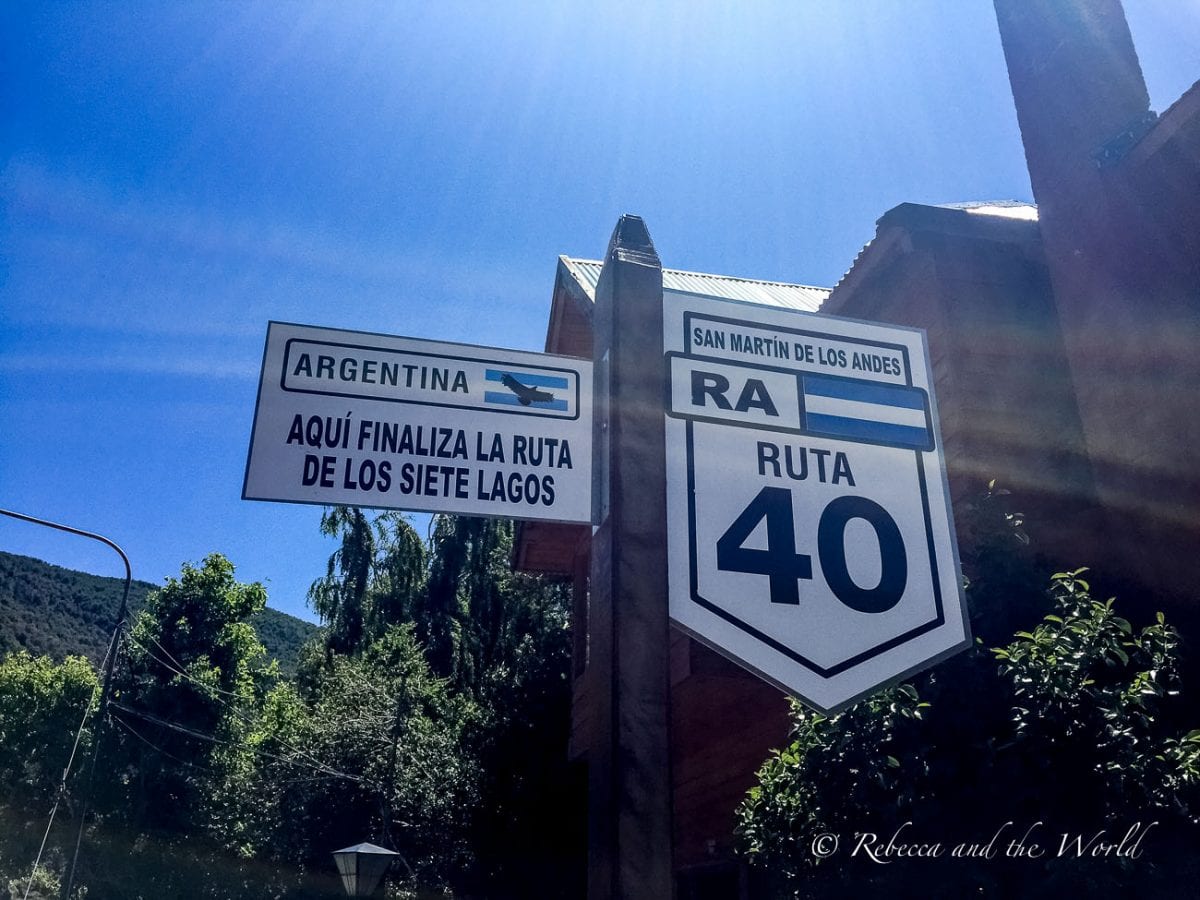 A street sign at a crossroad, with blue skies in the background. The sign on the left points to "Argentina" with a bird icon and below, text reading "AQUÍ FINALIZA LA RUTA DE LOS SIETE LAGOS" (which means "the Ruta de los Siete Lagos finishes here"). The sign on the right shows "SAN MARTIN DE LOS ANDES" above a large shield with "RA RUTA 40". The Ruta de los 7 Lagos is on the famous Ruta 40 in Argentina, the highway that stretches the length of the country.