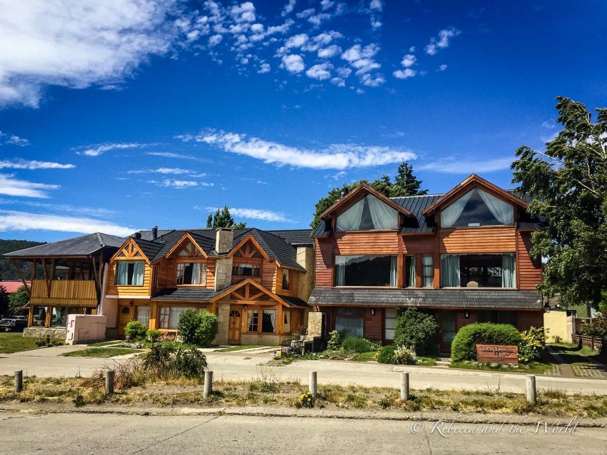 A view of a timber two-story house in an alpine style, with large windows and a gabled roof. The house is set against a backdrop of trees and a blue sky with scattered clouds. San Martin de los Andes in Argentina is a cute town to base yourself in to visit the 7 Lakes Route. 