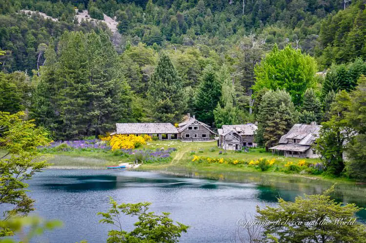 The best time to visit Argentina is spring when temperatures are milder and crowds fewer. Spring in Argentina is a great time to visit the Ruta de los Siete Lagos (Seven Lakes Route), seen in this image. Several stone and timber buildings sit in front of a lush green forest by the side of a lake. Yellow and purple flowers spring up between the grass.