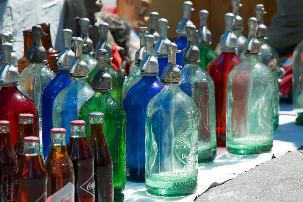 A collection of colorful vintage soda siphon bottles displayed on a table. The glass bottles come in shades of blue, green, and red, with metallic dispensing caps. These siphons were found at the San Telmo Market in Buenos Aires, Argentina.