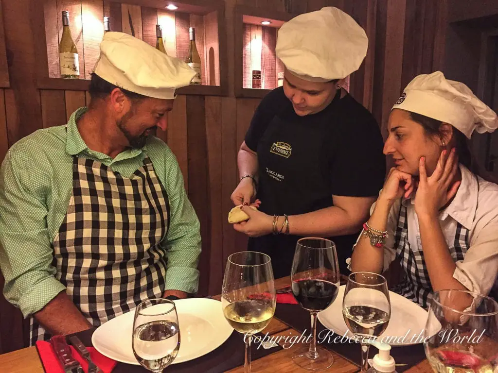 An indoor dining scene with three people wearing chef's hats. A man on the left wearing a green checkered shirt and apron is smiling while a woman in the center, in black attire, is demonstrating something with a piece of dough. Another woman on the right in stripes appears to be watching intently. They are seated at a table with wine glasses and a bottle. They are participating in The Argentine Experience, one of the best things to do in Buenos Aires.