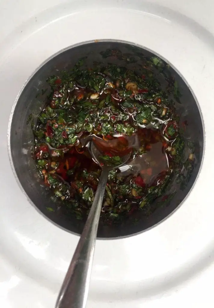 An overhead view of a red and green sauce called chimichurri in a white bowl with a spoon in the bowl. Chimichurri is a traditional Argentinian sauce usually served with steak.