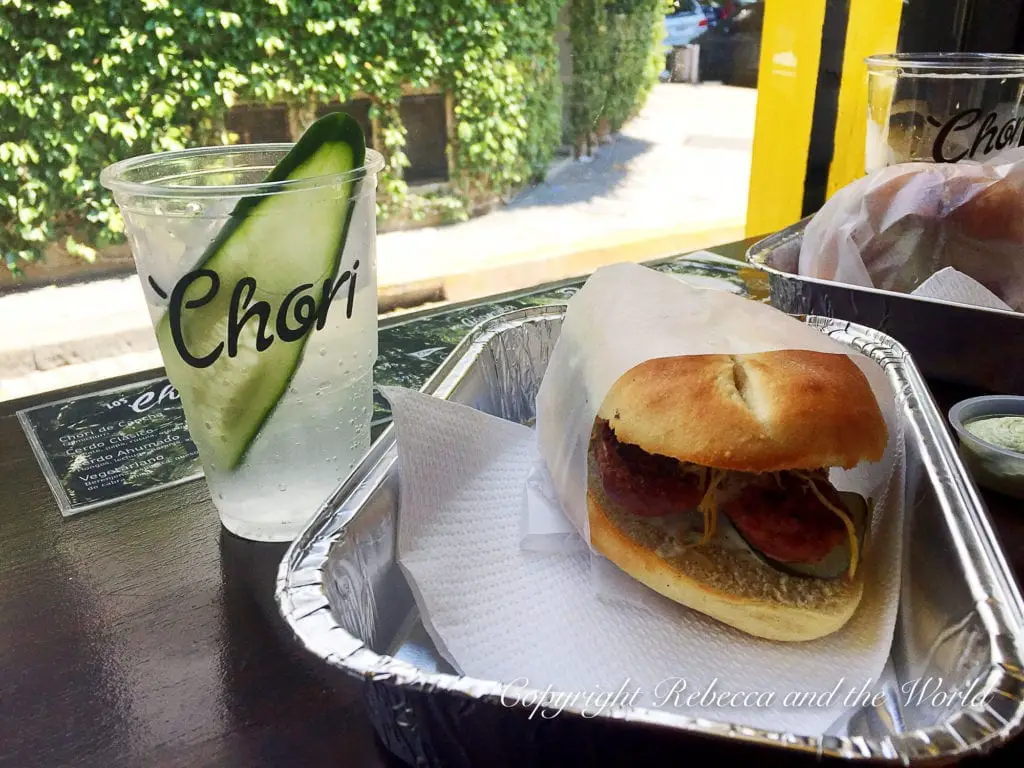A sandwich with chorizo on a white plate, a water glass with a cucumber slice, and a blurred menu in the background. Choripan is a great snack to try in Buenos Aires, Argentina - it's a sausage in bread smothered in chimichurri sauce and is one of the best eats in Buenos Aires.