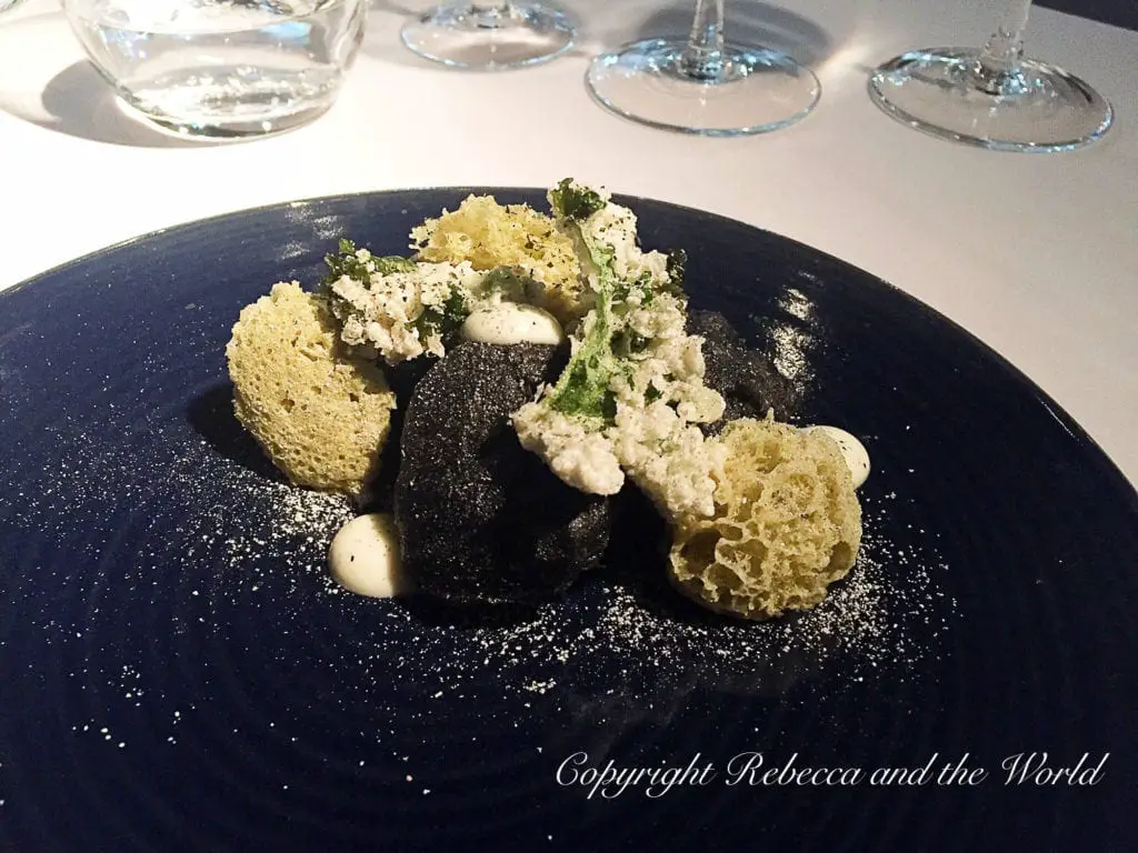 A dark blue plate with an artistic food presentation, featuring a black sauce, crumbs, and light-coloured foam. There are many wonderful fine dining restaurants in Buenos Aires - plan a special night out.