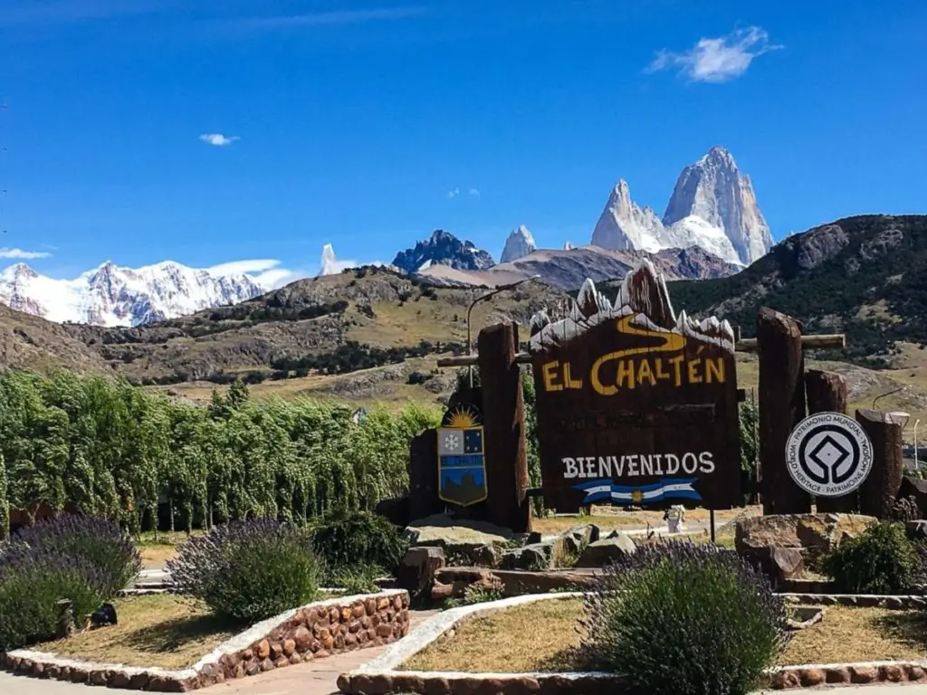 The entrance to El Chaltén, Argentina, featuring a large wooden sign with the town's name and "Bienvenidos" underneath, with a backdrop of towering, snow-capped mountains and a clear blue sky.