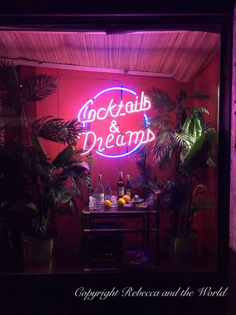 A neon sign reading "Cocktails & Dreams" above a bar setup with bottles, glasses, and fruits, set against a pink wall with potted plants. One of the best bars in Buenos Aires is Floreria Atlantico, a secret bar hidden underneath a florist.