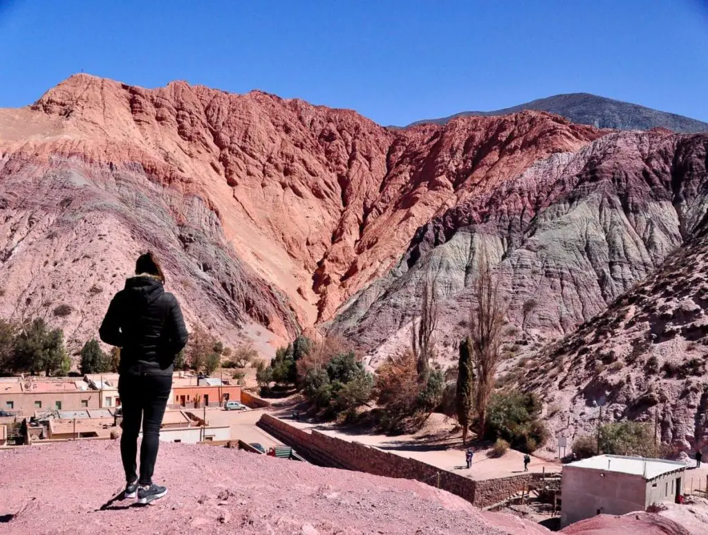 A woman (the author of this article) stands with their back to the camera, overlooking a dramatic landscape of multicolored mountain slopes in shades of red, purple, and gray - this is the Cerro de Siete Colores (Hill of 7 Colours). A small village with terracotta rooftops is nestled at the mountain's base beneath a clear blue sky.