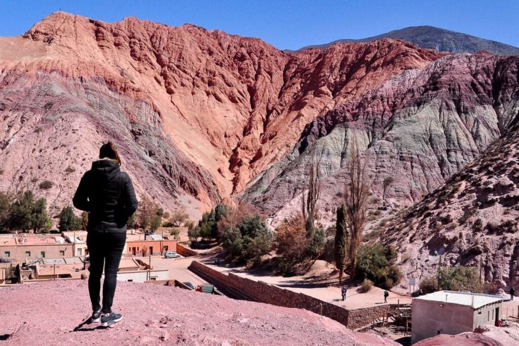 A woman (the author of this article) stands with their back to the camera, overlooking a dramatic landscape of multicolored mountain slopes in shades of red, purple, and gray - this is the Cerro de Siete Colores (Hill of 7 Colours). A small village with terracotta rooftops is nestled at the mountain's base beneath a clear blue sky.