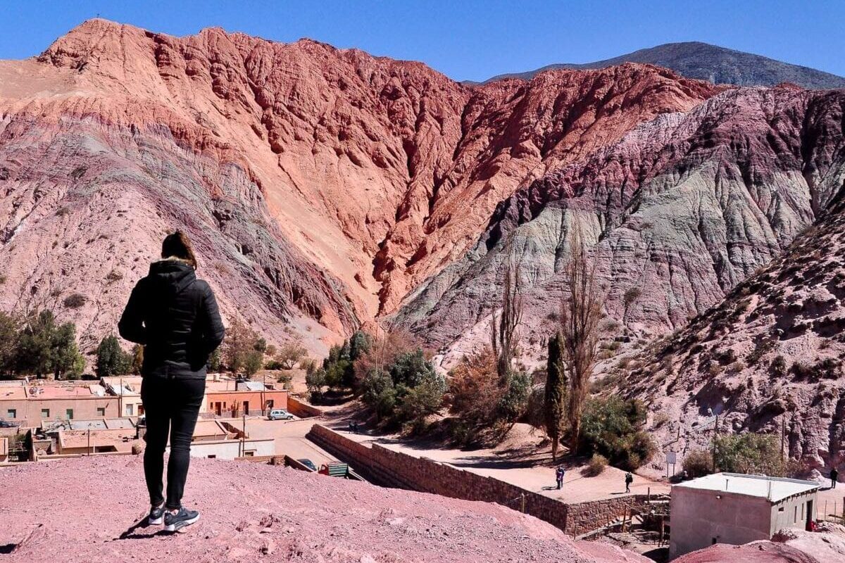A woman (the author of this article) stands with their back to the camera, overlooking a dramatic landscape of multicolored mountain slopes in shades of red, purple, and gray - this is the Cerro de Siete Colores (Hill of 7 Colours). A small village with terracotta rooftops is nestled at the mountain's base beneath a clear blue sky.