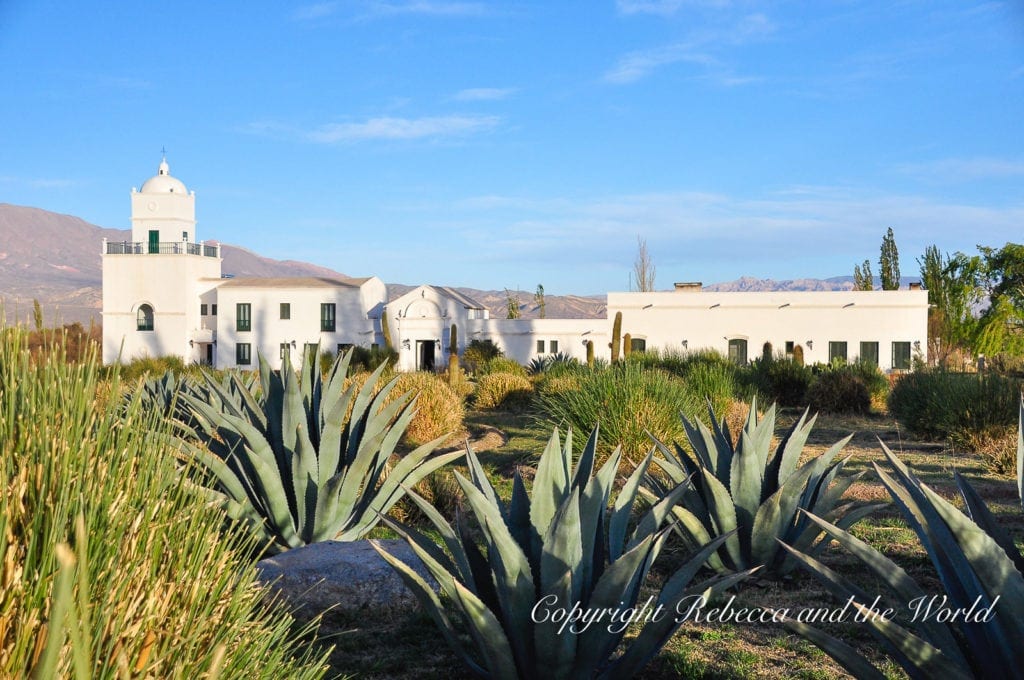 A white colonial-style building with a bell tower surrounded by agave plants, against a backdrop of mountains under a clear blue sky in North Argentina. Cachi is a charming village in north Argentina that's worth a day or two.