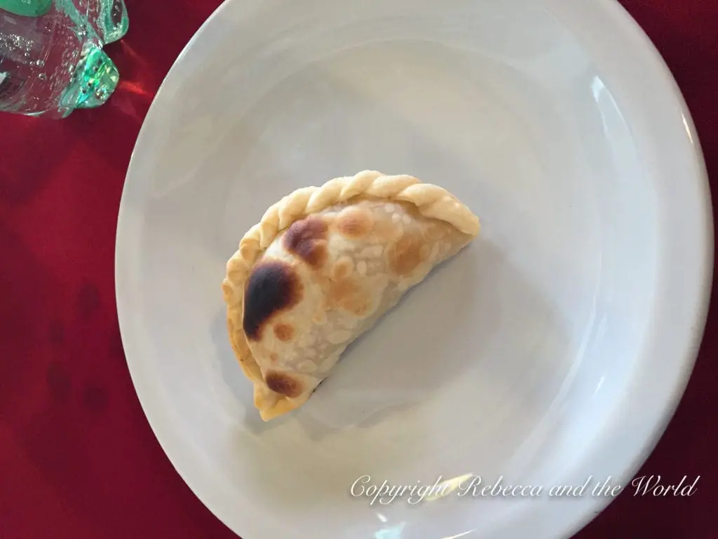 A single golden-brown empanada with a fluted edge is centered on a white plate, with a portion of a green glass visible in the upper left corner and set against a red background. You must try empanadas saltenas when you visit northern Argentina - they're delicious!