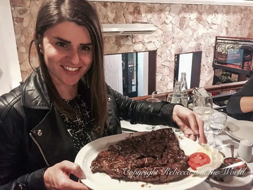 A smiling woman - the author of this article - sitting at a restaurant table, holding a large plate with a grilled steak, typical Argentinian food. In Salta - like most of Argentina - you can eat steaks the size of your head!