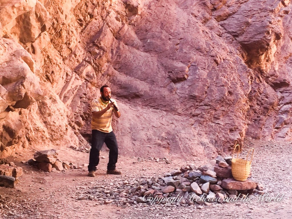 A person in a yellow shirt playing a flute in a rocky desert setting with a woven basket in the foreground and red rock cliffs in the background in North Argentina. The Quebrada de las Conchas is home to the Garganta del Diablo, a huge cavern where performers often play.
