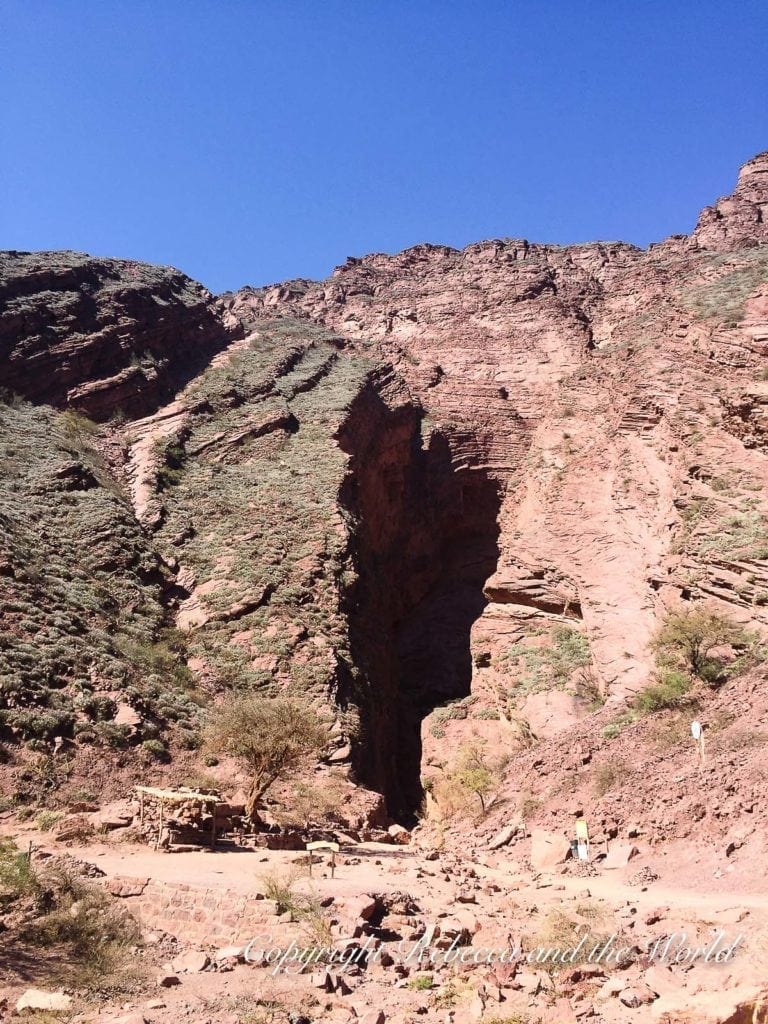 A large cave opening in a red rock mountain face with a small figure standing at the entrance for scale, under a clear blue sky in North Argentina. The final day of the north Argentina road trip will see you driving past amazing rock formations and landscapes.