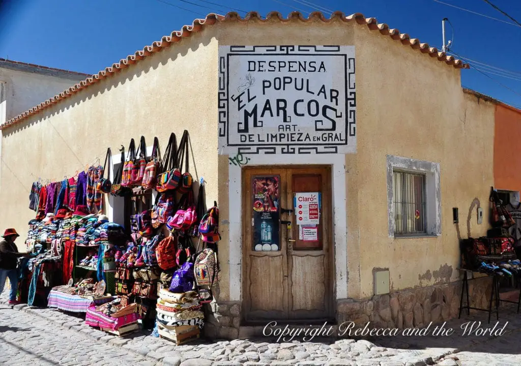 A colorful storefront with traditional textiles and handicrafts displayed outside. The sign above reads "Despensa Popular Marcos" in bold letters, indicating a general store. The store is in North Argentina.