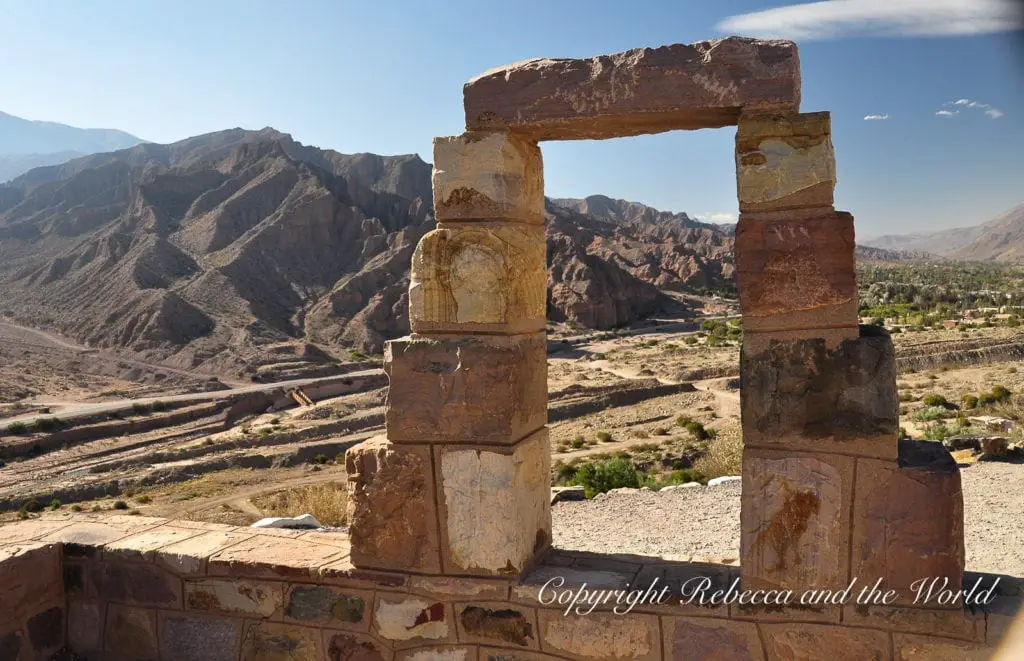 A stone archway framing a picturesque view of eroded hills and a valley with sparse greenery under a blue sky in North Argentina. In Tilcara in the north of Argentina, visit the pre-Incan fort of Pucara.