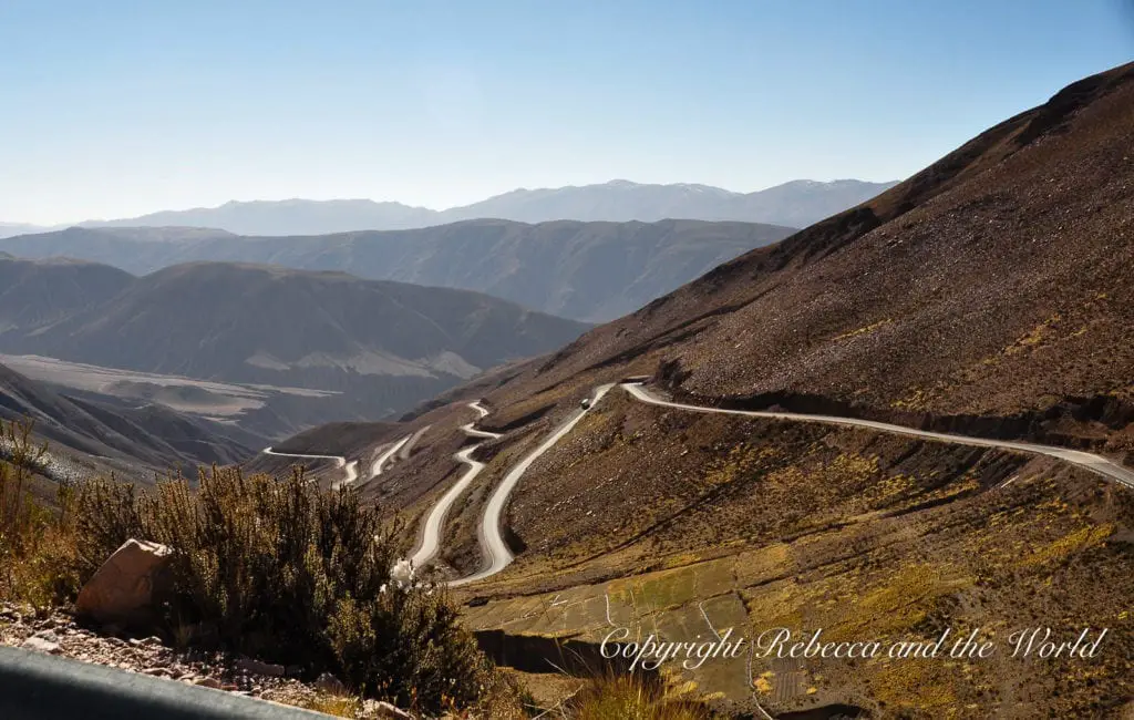A winding mountain road in Northwest Argentina cutting through a dry, hilly landscape with distant mountain ranges under a clear blue sky.