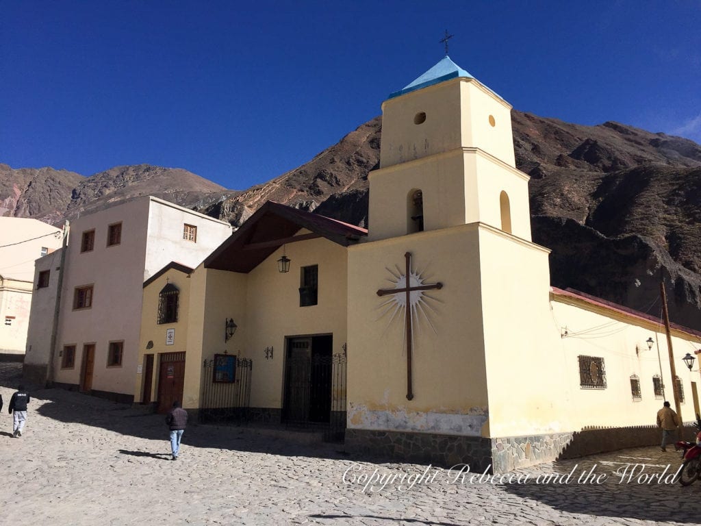 A bright yellow church with a simple facade and a bell tower in a cobblestone plaza, set against a clear blue sky in North Argentina. The town of Iruya is one of the most interesting places to visit in Argentina.
