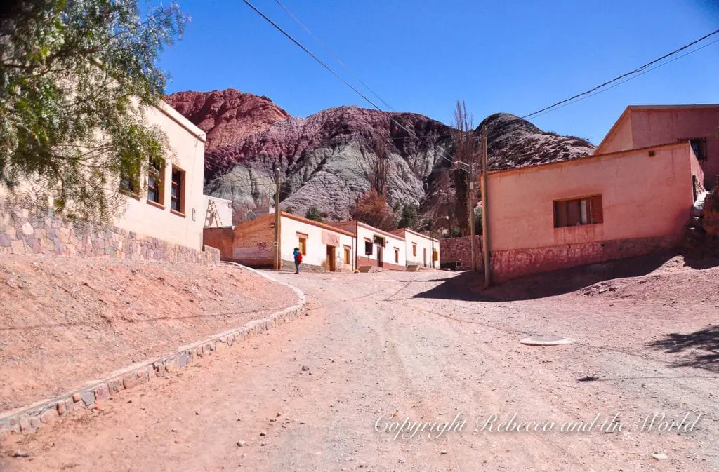 A dirt road winds its way between low adobe buildings. In the background is the Cerro de Siete Colores (Hill of 7 Colours) in Purmamarca, Argentina, with layers of different coloured sediments.