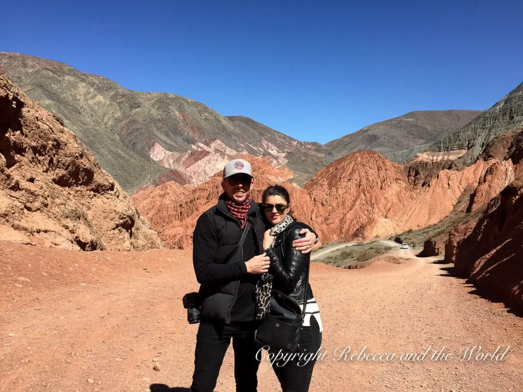 The author of this blog post with her husband in Purmamarca, North Argentina - a fabulous honeymoon destination in Argentina. A man and woman, both in black clothing, standing on a dirt road, surrounded by colourful mountains.