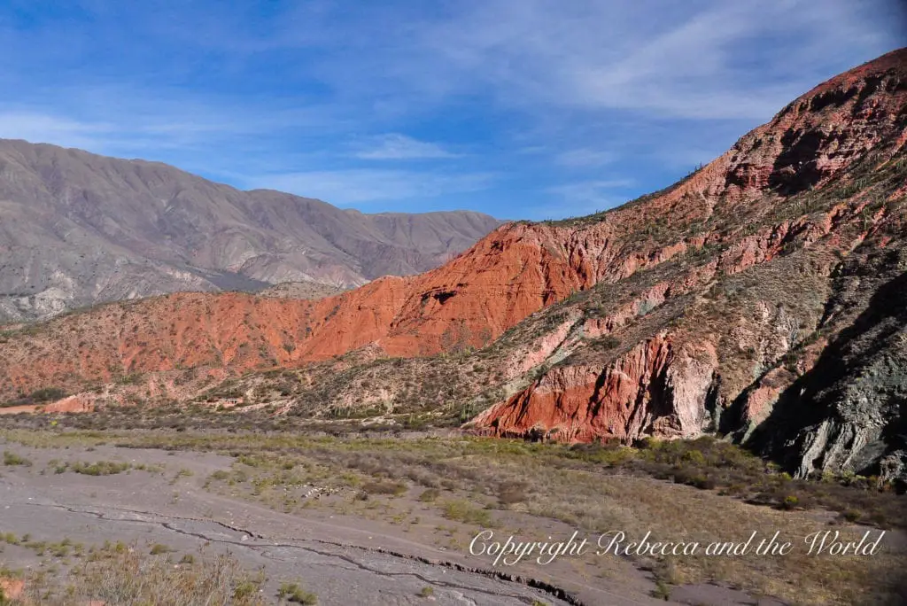 A striking landscape showing red rock formations with contrasting layers of colour against a backdrop of green vegetation and a clear sky. This is Northern Argentina.