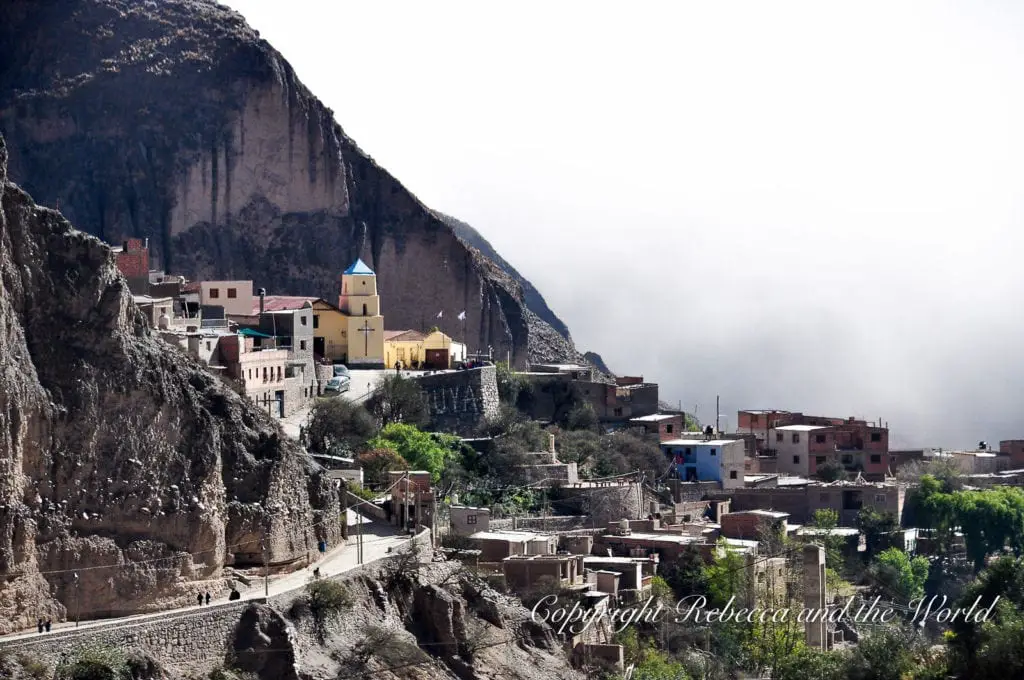A breathtaking view of the secluded village of Iruya, perched high on a mountainside. The colourful houses and a bright yellow church stand out against the rugged cliffs, shrouded partially in mist, giving the scene a mystical, timeless atmosphere.
