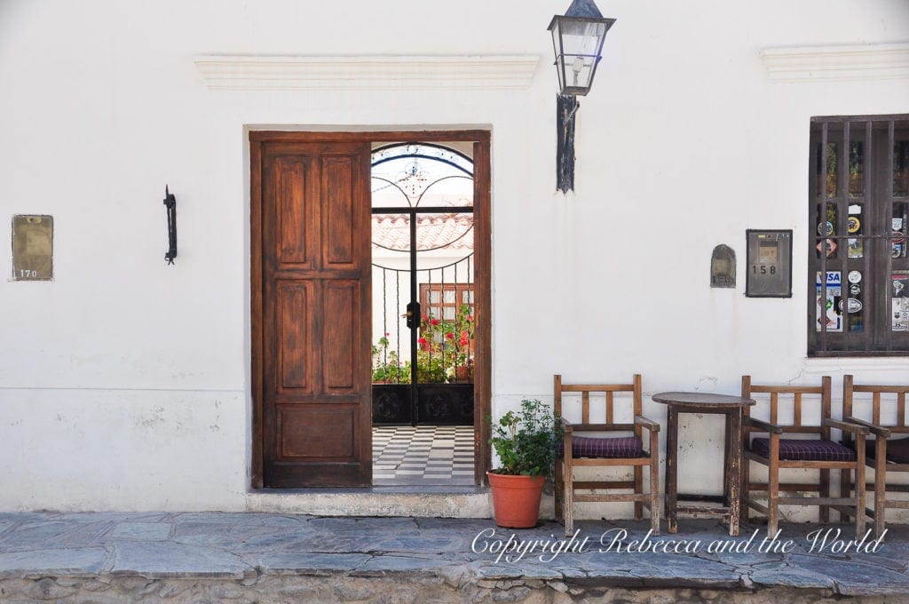 A cozy street corner in North Argentina featuring a wooden doorway opening to a garden, a lantern on the wall, and a couple of chairs with a table on the cobblestone sidewalk. Cachi is a charming village in north Argentina that's worth a day or two.