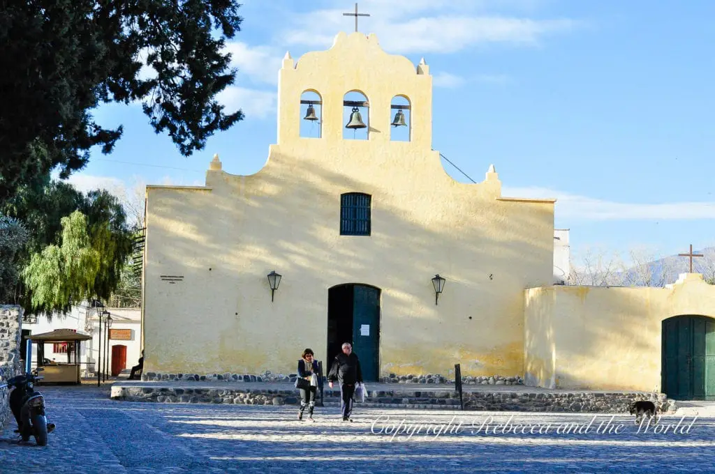 A simple, traditional church with a bell tower and three bells visible against a clear sky in a cobbled square, with a couple of people walking out of the doorway in North Argentina. Cachi is a charming village in north Argentina that's worth a day or two.