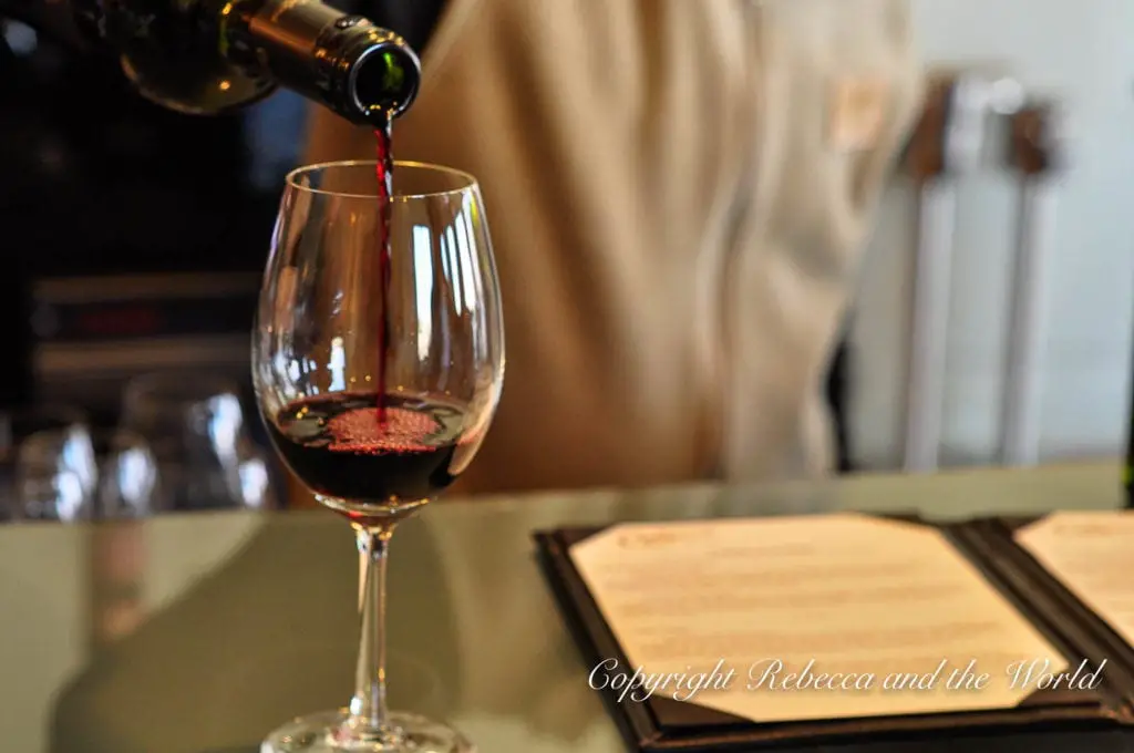 A close-up of red wine being poured into a clear glass, capturing the rich color and smooth flow of the wine, set against a blurred background.