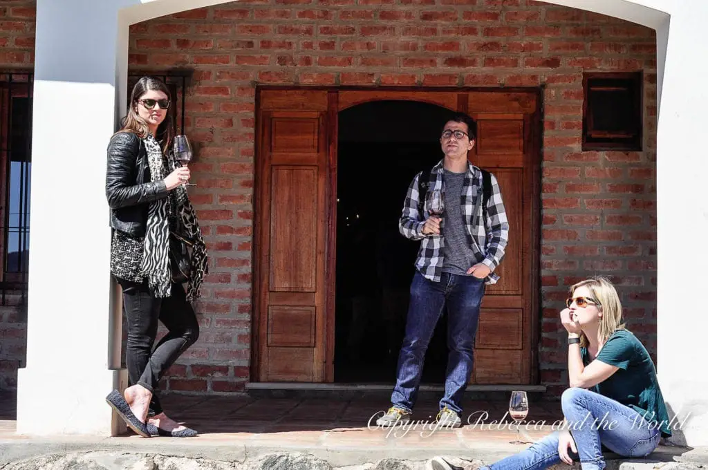 Three people - the author and her friends - standing and sitting outside a brick building with wooden doors, casually enjoying glasses of wine in the sun.