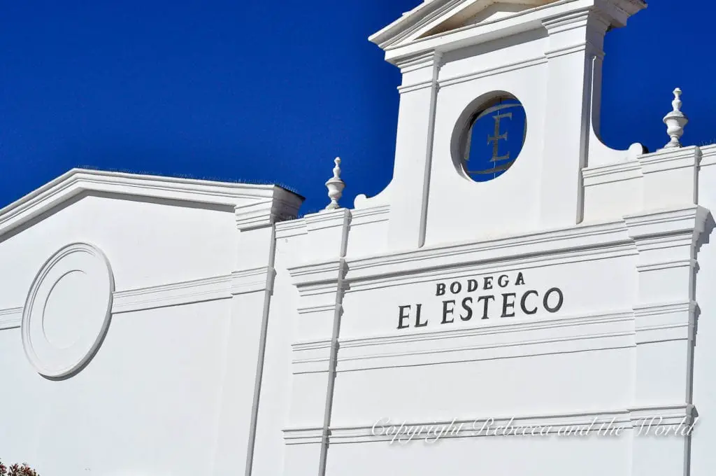 The exterior of Bodega El Esteco, showing a white building with a classical design, elegant trim, and the winery's name and initial in bold lettering.