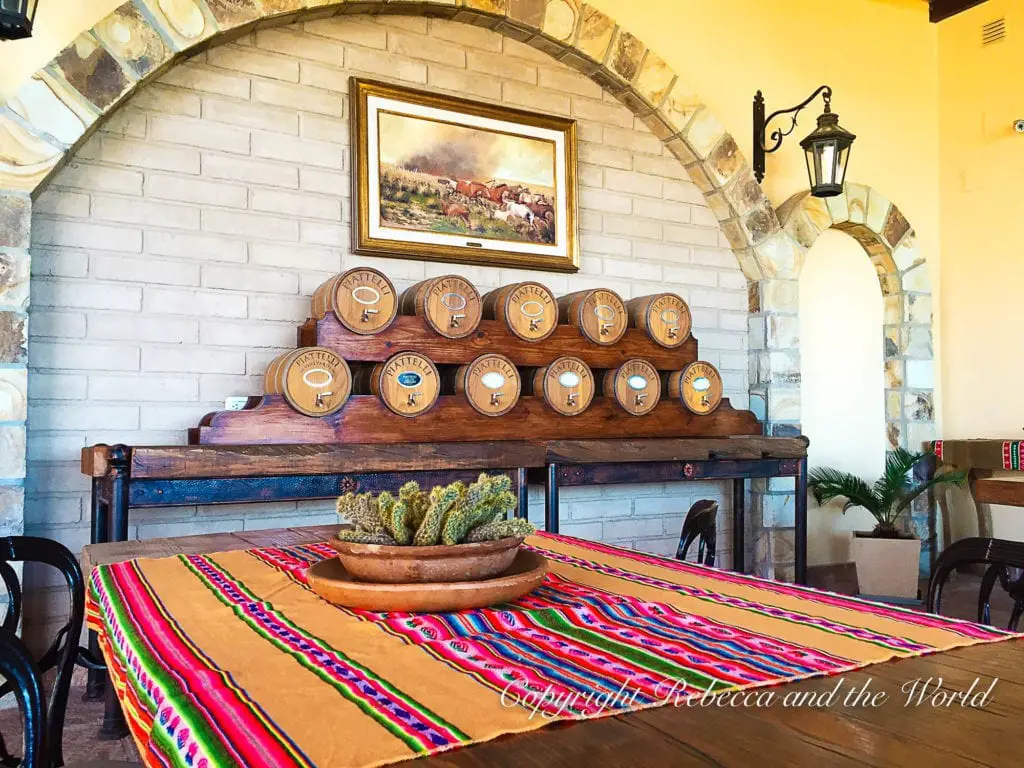 An interior rustic setting with a table covered in a colorful striped cloth, wooden barrels marked with wine varieties on a shelf, and a brick archway in the background. Piattelli is one of the best wineries in Cafayate.