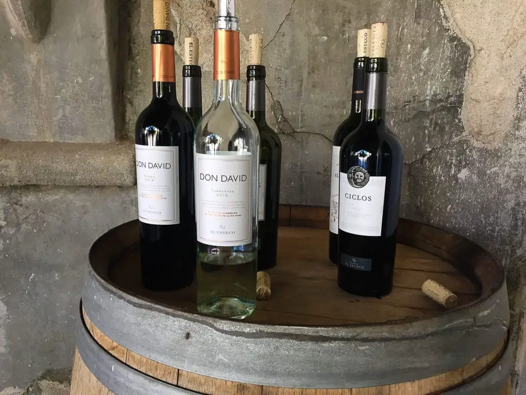 Three bottles of wine, two red and one white, placed on a wooden barrel. The labels show the brand "Don David" for the red wines and "Ciclos" for the white. A stone wall provides the backdrop. Wine is a great Argentina souvenir to bring home with you.