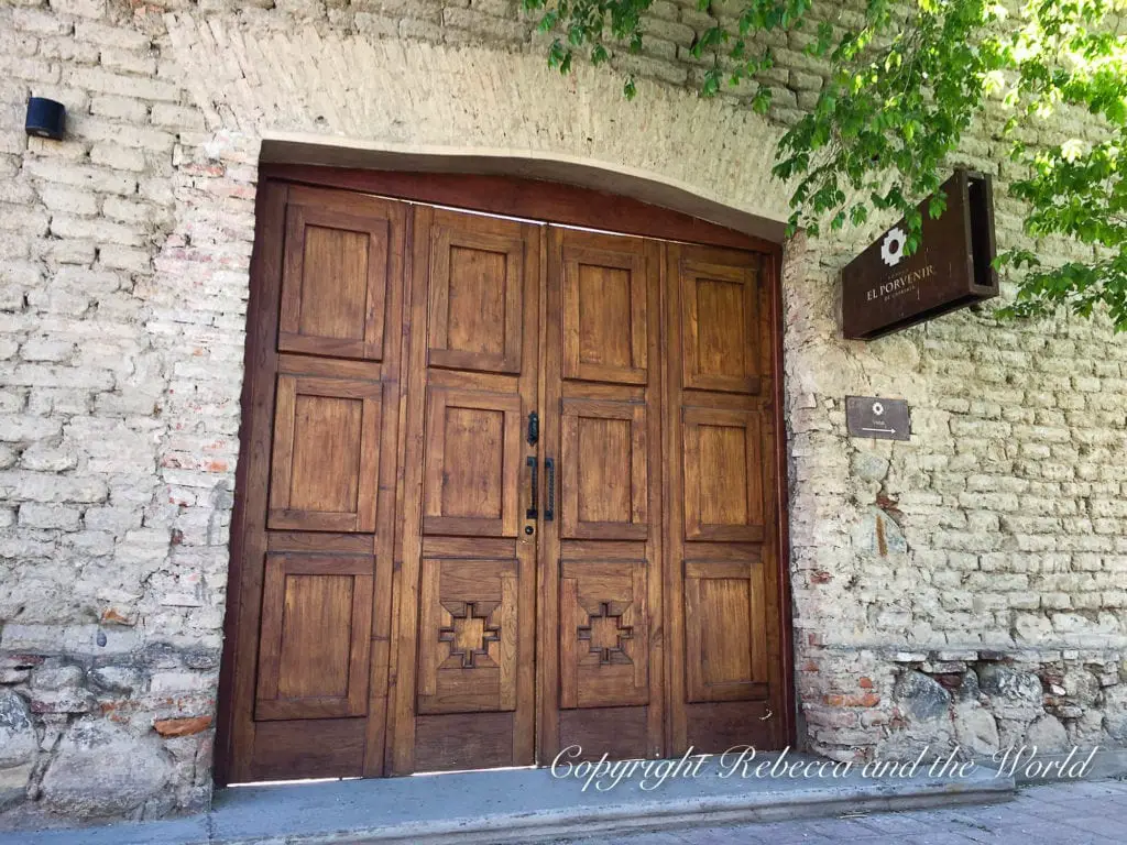 A traditional, large wooden door entrance to El Porvenir winery, with a discreet sign next to a stone wall covered in green foliage.