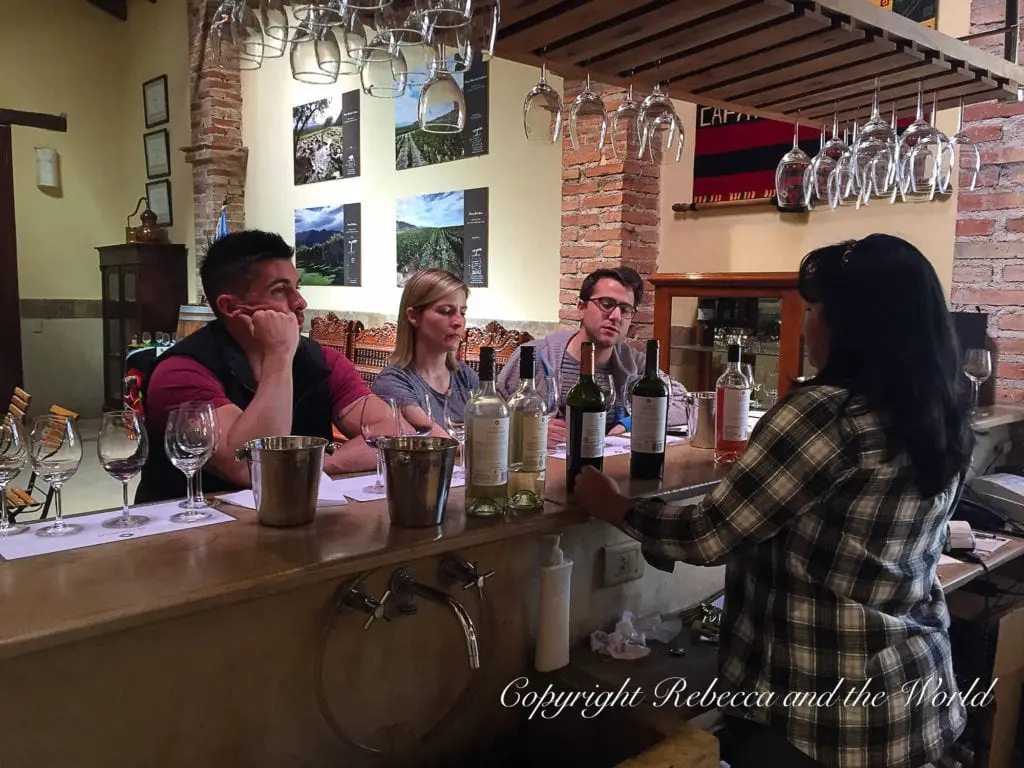 An informal wine tasting in progress, with a group of people - the author's husband and her friends - sitting at a bar while a server pours wine, in a cozy setting with brick walls and hanging wine glasses.