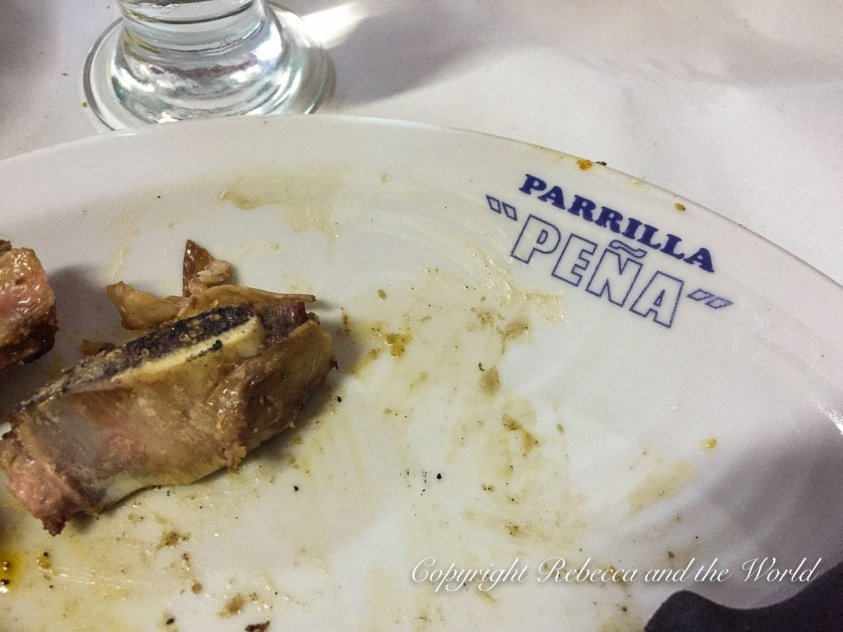 Close-up of a partially eaten meal on a white plate with the text "PARRILLA 'PEÑA'" in blue letters near the rim. Visible are a few pieces of meat with some fat and gristle, and remnants of a sauce or seasoning. The background is unfocused, with a hint of a glass object on the left edge. This is one of the best steakhouses in Buenos Aires, Parrilla Pena.