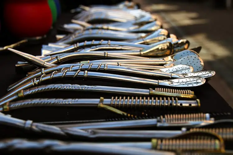 A collection of silver bombillas (straws) laid out on a dark surface. The bombillas have ornate handles and are arranged in a neat row. The silver bombilla is a gorgeous thing to buy in Buenos Aires.