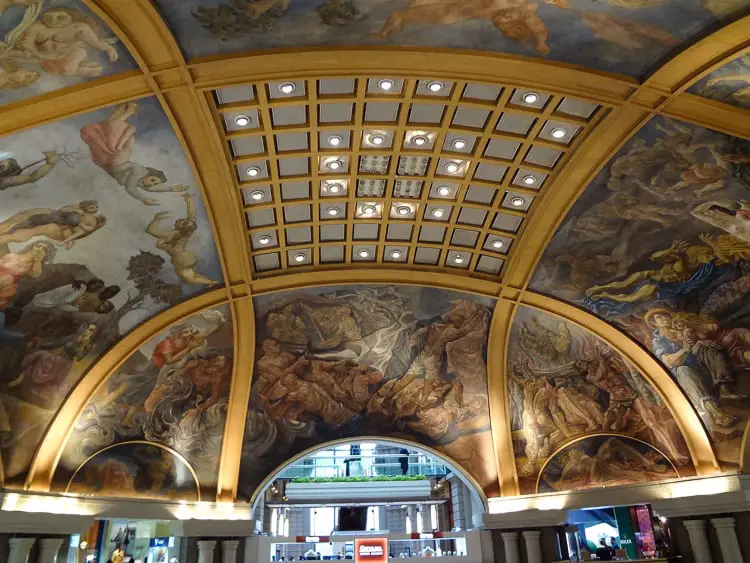 A ceiling with elaborate murals depicting classical figures and scenes, framed by a golden arch. The architecture features a grid of illuminated panels. Galerias Pacifico is one of the most famous Buenos Aires shopping malls.