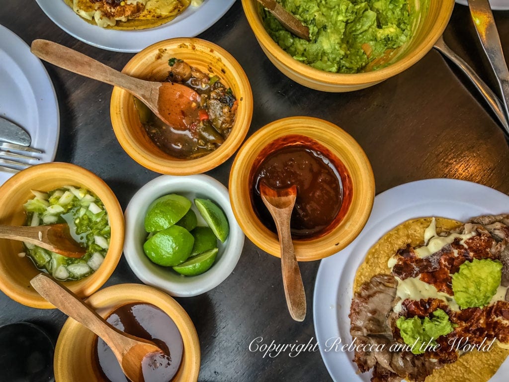 An overhead view of a table with various Mexican salsas and condiments in clay bowls, including chopped onions, limes, and a plate of tacos with guacamole.