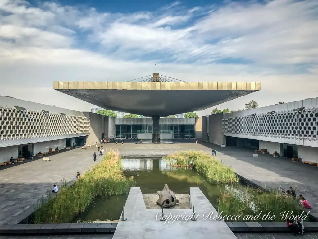 The expansive, modern courtyard of the National Museum of Anthropology in Mexico City, featuring a large square reflecting pool and a striking, cantilevered roof structure.