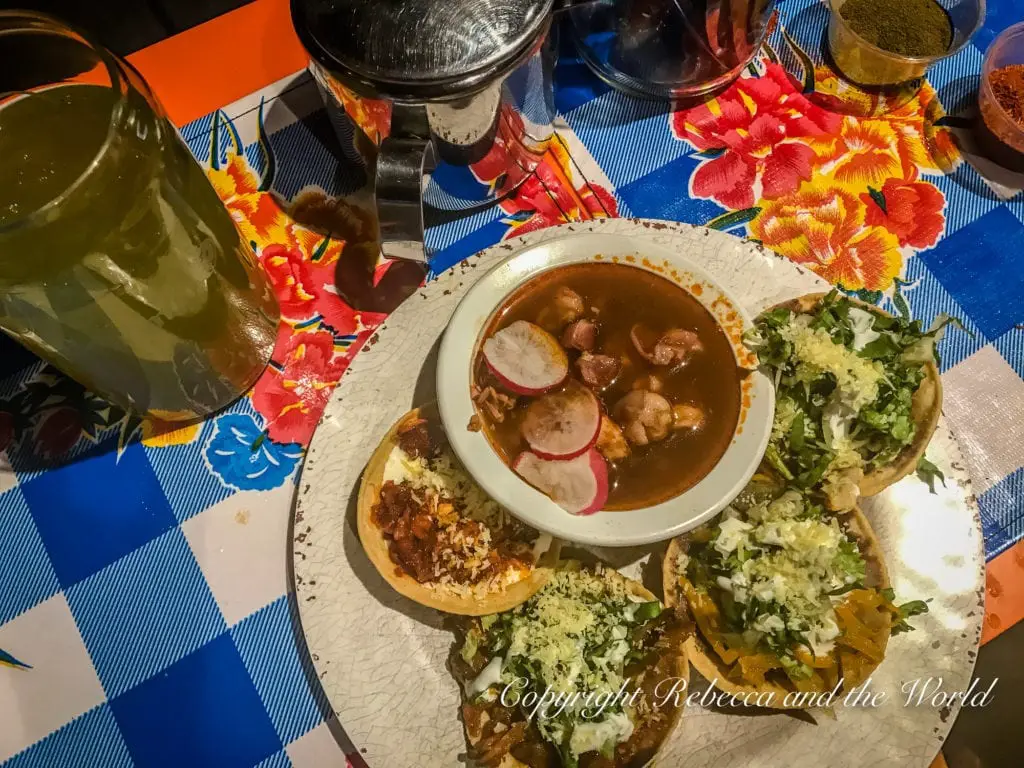 A plate of classic Mexican pozole, a traditional soup or stew, accompanied by tostadas topped with lettuce and cheese, presented on a table with colourful tablecloth.