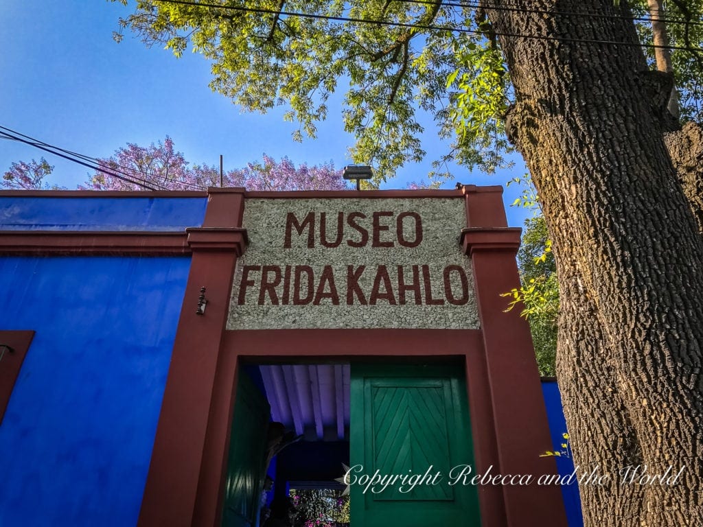 The entrance to the Museo Frida Kahlo, with a bold sign above the door, set against the museum's iconic blue walls, with trees and a blue sky in the background.