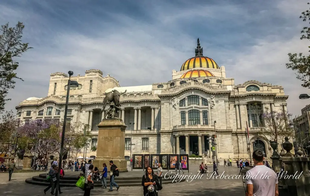 The grand Palacio de Bellas Artes in Mexico City, with its distinctive yellow and orange dome, captured on a sunny day with pedestrians and a statue in the foreground.