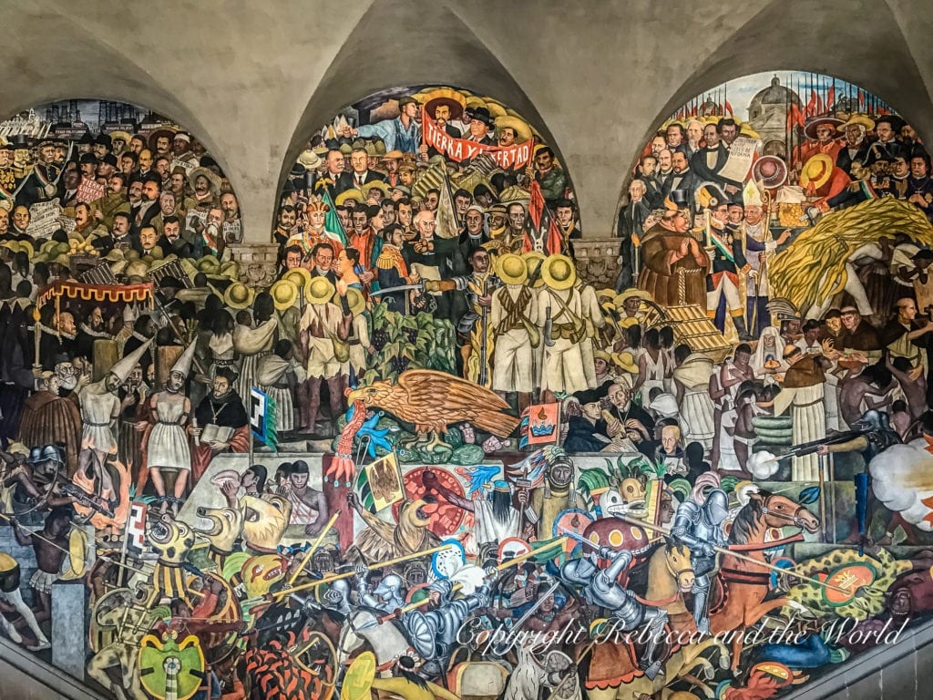 A detailed mural by Mexican artist Diego Rivera, filled with dense crowds of people from different eras of Mexican history, including soldiers, labourers, and activists, with symbolic imagery interwoven throughout.